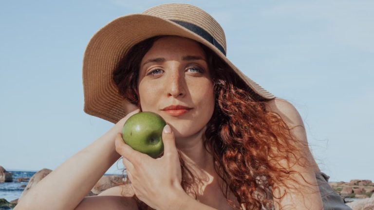 A relaxed woman with a hat holds a green apple, enjoying a sunny beach day.