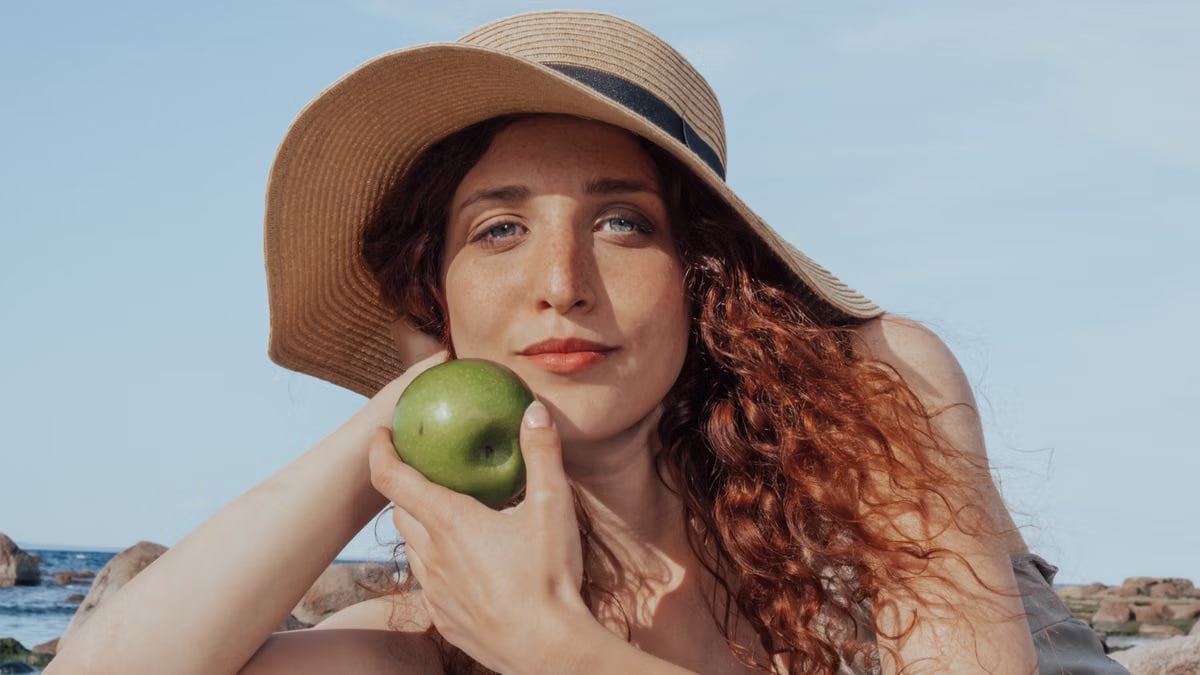 A relaxed woman with a hat holds a green apple, enjoying a sunny beach day.